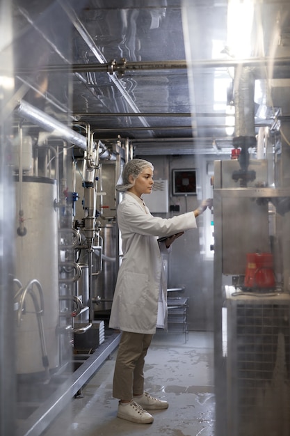 Verticaal portret over de volledige lengte van een jonge vrouw die machine-eenheden bedient terwijl ze in een industriële voedselfabriek werkt