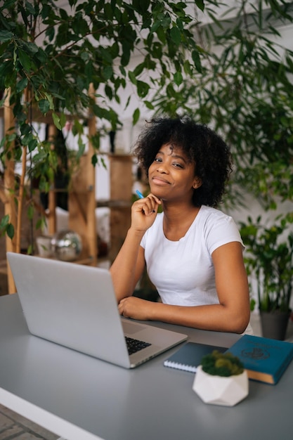 Verticaal portret van aantrekkelijke Afro-Amerikaanse jonge vrouw met pen in de hand glimlachend kijkend naar camera zittend aan tafel in lichte kantoor aan huis kamer