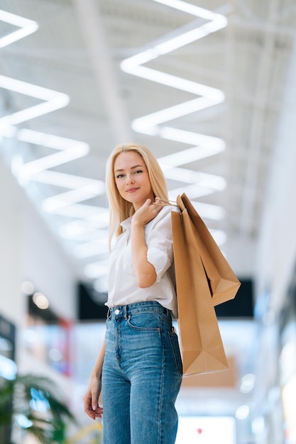 Verticaal portret van aantrekkelijke blonde jonge vrouw met papieren boodschappentassen met aankopen