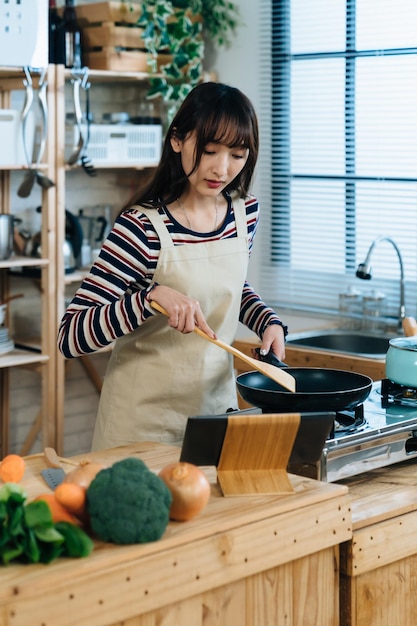 verticaal portret van aziatische nieuwe vrouw kijkt naar online instructievideo op tabletcomputer om te leren lunchen terwijl ze thuis een koekenpan in de keuken gebruikt.