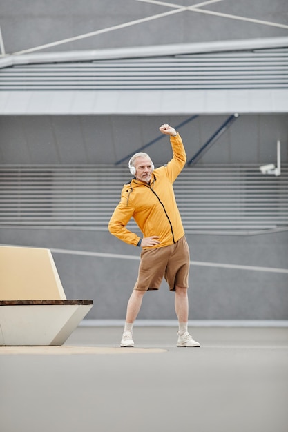 Verticaal portret van de volledige lengte van een sportieve volwassen man die zich buiten uitstrekt met een koptelefoon in stedelijke omgeving