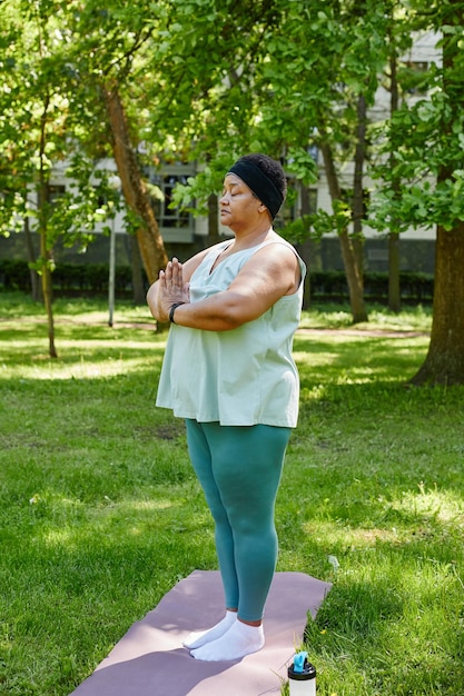 Verticaal portret van de volledige lengte van een zwarte vrouw met overgewicht die buiten yoga doet in een groen park en medita