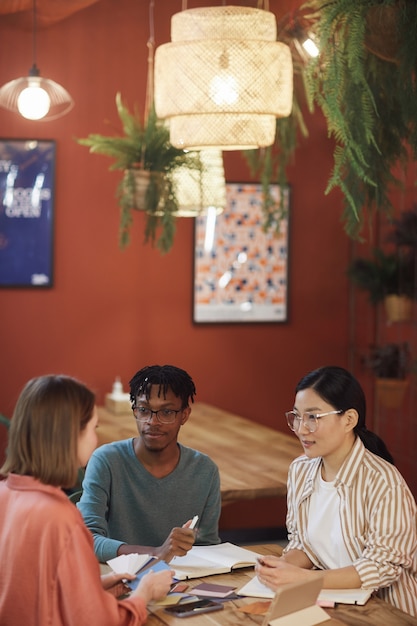 Verticaal portret van drie jonge mensen die aan project werken zittend aan tafel in café tegen rode muur