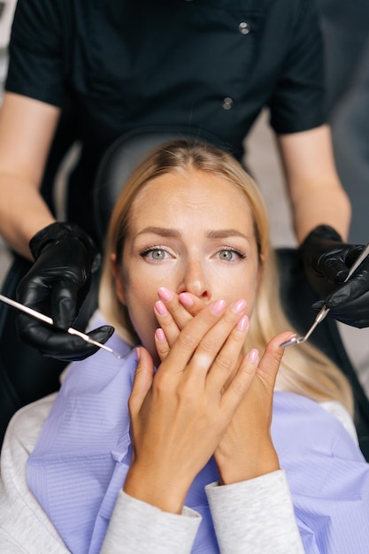 Verticaal portret van een angstige blonde vrouwelijke patiënt die met haar handen de mond sluit en het onderzoeken van de tanden verhindert omdat ze uit angst naar de camera kijkt met een angstige uitdrukking