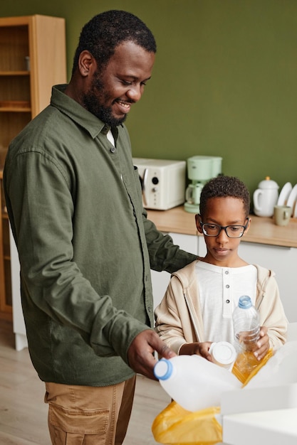 Verticaal portret van een gelukkige Afro-Amerikaanse vader en zoon die plastic thuis in recyclingbakken zetten