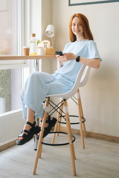 Verticaal portret van een gelukkige jonge vrouw in casual kleding met een mobiele telefoon aan het bureau bij het raam in een gezellig licht café. Mooie roodharige blanke dame met vrijetijdsbesteding in de coffeeshop.