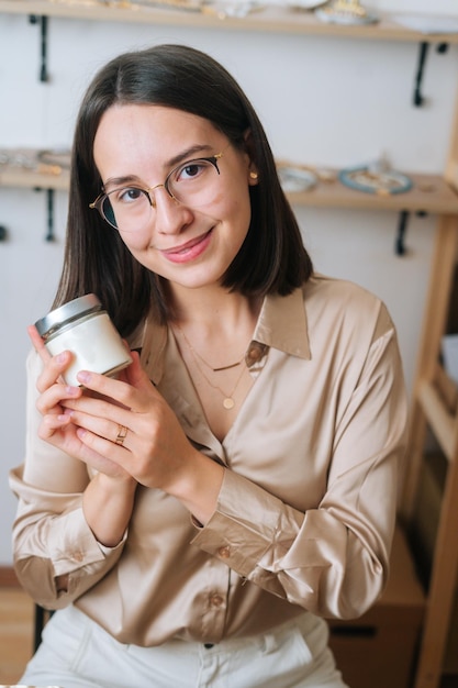 Verticaal portret van een glimlachende jonge vrouw in een bril met een kleine transparante glazen pot met handgemaakte geurkaars die naar de camera kijkt