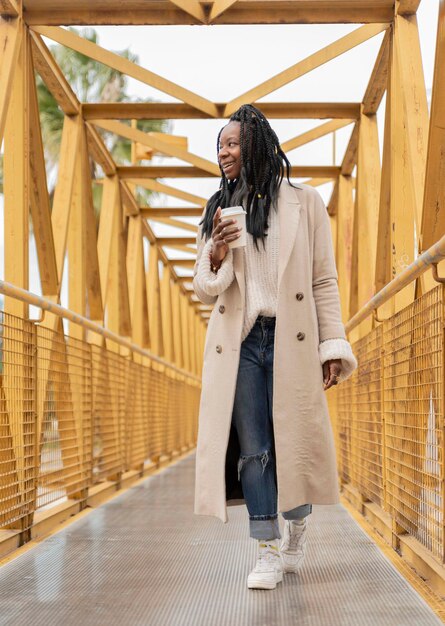 Verticaal portret van een jonge Afro-Amerikaanse vrouw met vlechten die over een gele brug loopt met een kopje koffie