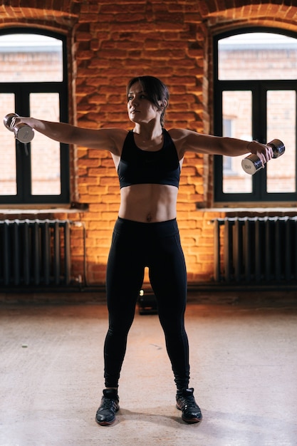 Verticaal portret van een jonge atletische vrouw met een mooi sterk lichaam die activewear draagt met halters op ingetrokken armen. Kaukasische vrouwelijke fitnesstraining uit te oefenen in een donkere sportschool.