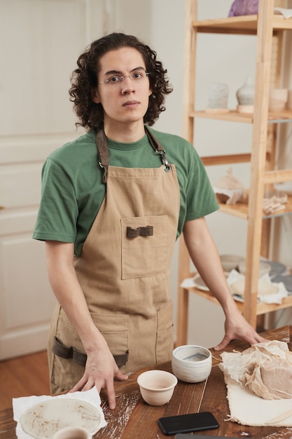 Verticaal portret van een jonge mannelijke ambachtsman die naar de camera kijkt terwijl hij bij het werkstation staat in de pottenbakkerij