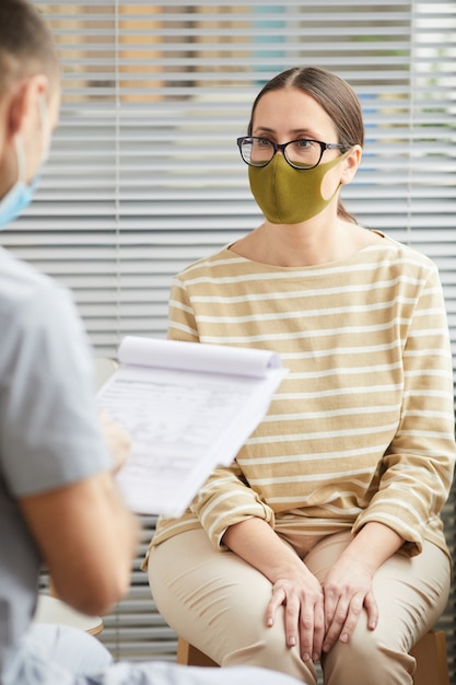 Verticaal portret van een jonge vrouw die een masker draagt en naar de dokter luistert tijdens het wachten in de rij bij de medische kliniek