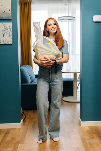 Verticaal portret van een mooie jonge vrouw die een boodschappentas vasthoudt en glimlacht terwijl ze naar de camera kijkt die op de stoep van het appartement staat