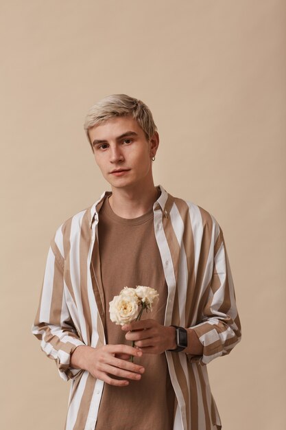 Verticaal portret van een vrouwelijke blonde man die bloemen vasthoudt en naar de camera kijkt terwijl hij tegen een neutrale beige achtergrond in de studio staat