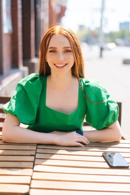 Verticaal portret van gelukkig lachende jonge vrouw zittend aan tafel op terras van café in zonnige zomerdag en kijken naar camera, onscherpe achtergrond, selectieve focus.