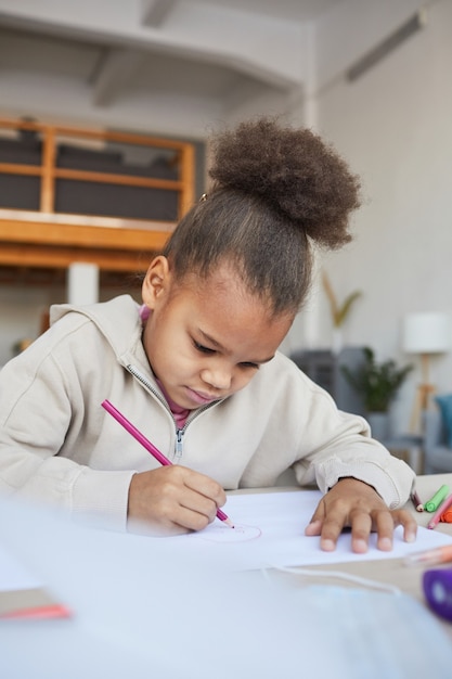 Verticaal portret van schattig afrikaans-amerikaans meisje dat foto's maakt terwijl ze aan tafel zit in een gezellig interieur, kopieer ruimte