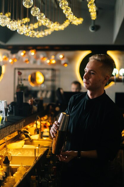 Verticaal portret van serieuze barman die ingrediënten van verse cocktail mengt door shaker te schudden die achter de toog staat, op onscherpe achtergrond van restaurant met donker interieur, selectieve focus