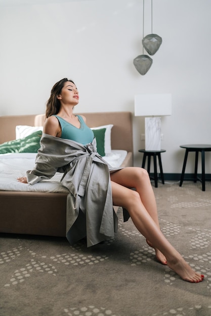 Verticaal portret van verleidelijke brunette vrouw in onthullend gewaad en beha zittend op bed in luxe slaapkamer in sensuele erotische houding met gesloten ogen