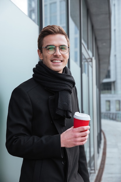Verticaal portret van zakenman in glazen en warme kleren die zijwaarts staan en buiten koffie drinken.
