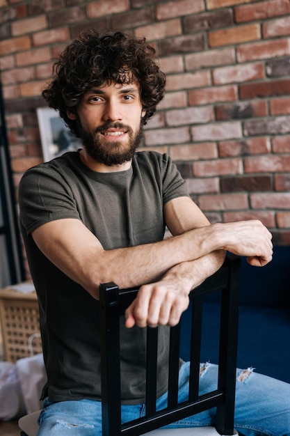 Verticaal portret van zelfverzekerde glimlachende jongeman die naar de camera kijkt terwijl hij op een stoel zit en zich vasthoudt aan chairbac in een gezellige woonkamer met bakstenen muur