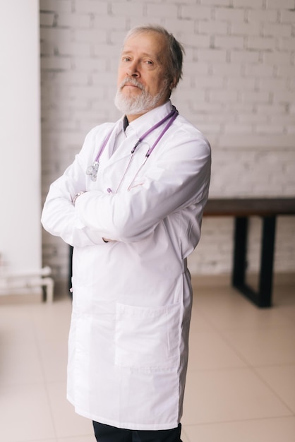 Verticaal portret van zelfverzekerde volwassen man arts met witte laboratoriumjas en stethoscoop poseren met gekruiste armen in het ziekenhuis. Professionele arts met witte baard en grijs haar op de werkplek.