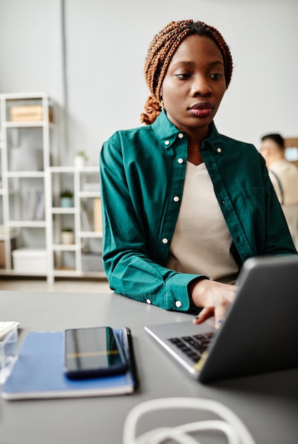 Verticaal portret van zwarte jonge vrouw die laptop gebruikt terwijl ze aan het bureau werkt in softwareontwikkelingsst