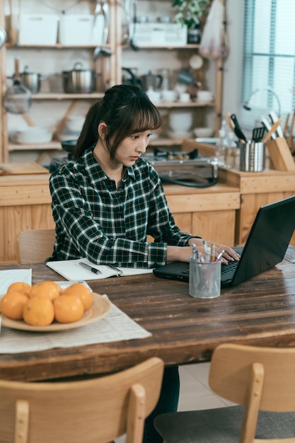 verticaal schot Koreaanse vrouwelijke student schrijft essay op laptop. Aziatisch meisje zit aan de eettafel en kijkt met volle aandacht naar het pc-scherm.