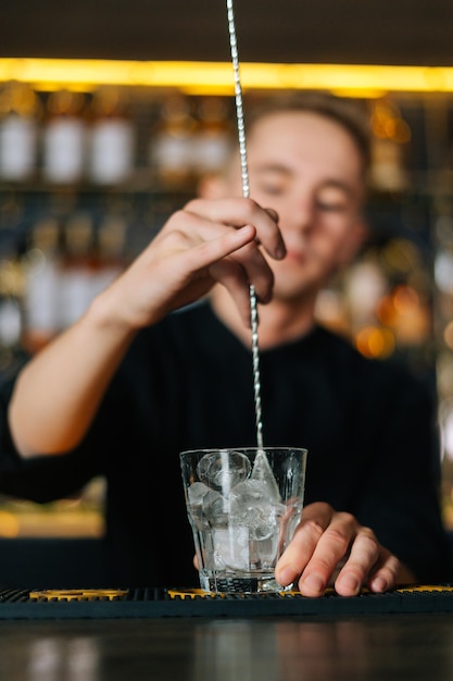 Verticaal schot van barman die ijsblokjes in glas roert met behulp van een lange barlepel die op de selectieve focus van de bar staat