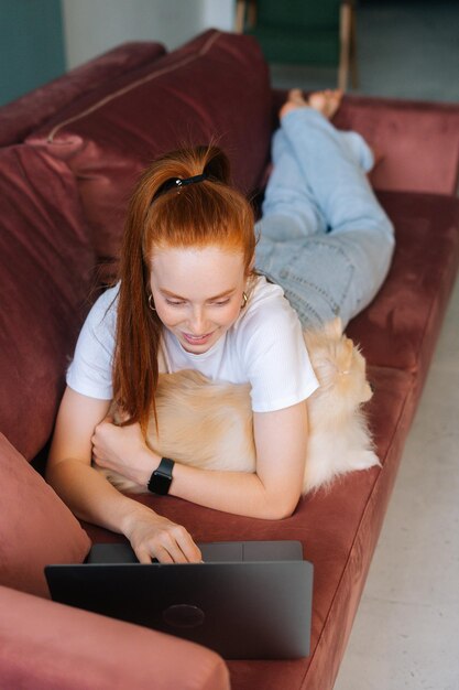 Verticaal schot van een gelukkige jonge vrouw die op een comfortabele bank ligt met een witte, mooie Spitz-hond en een laptop gebruikt in de woonkamer. Gelukkig roodharige vrouw tijd doorbrengen samen met schattige kleine hondje thuis