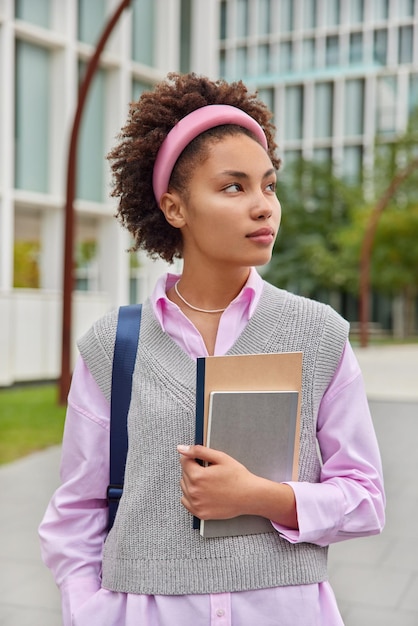 Verticaal schot van gekrulde vrouwelijke student gekleed in nette kleding draagt notebooks retourneert van lezingen gericht weg poses buitenshuis tegen onscherpe achtergrond Jeugd studeren en lifestyle concept