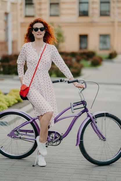 Verticaal schot van mooi roodharig vrouwelijk model heeft rust na lange afstand op de fiets te hebben afgelegd, draagt een zonnebril, zomerjurk en witte sneakers, geniet van sport en het verkennen van stadsstraten