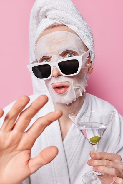 Foto verticaal schot van onder de indruk volwassen man houdt hand naar camera voelt verbaasd past schoonheid gezichtsmasker toe draagt zonnebril badjas en handdoek houdt glas cocktail geïsoleerd op roze achtergrond