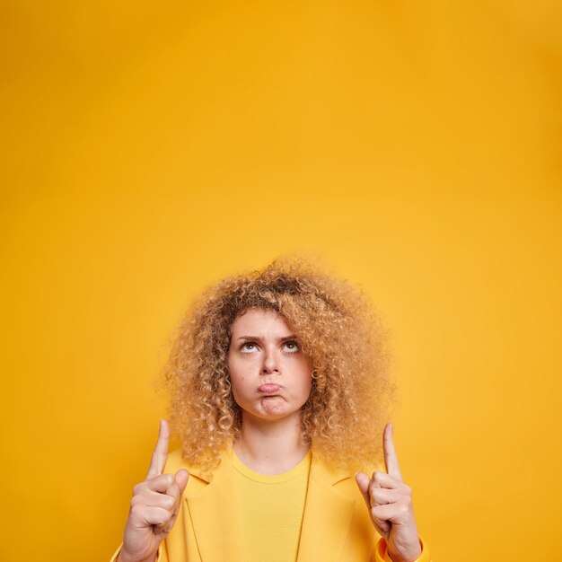 Verticaal schot van ontevreden boze jonge vrouw wijst boven portemonnees lippen houdt niet van iets dat formeel gekleed is geïsoleerd over levendige gele muur heeft ontevredenheid gezichtsuitdrukking. Advertentie