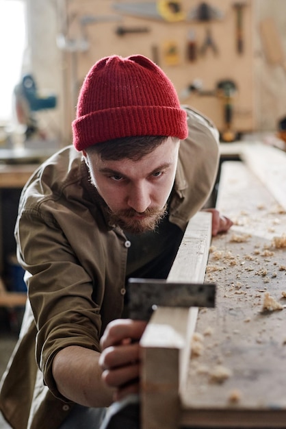 Foto verticaal schot van serieuze jonge mannelijke timmerman die stalen vierkant gebruikt bij het werken met houten plank in woo