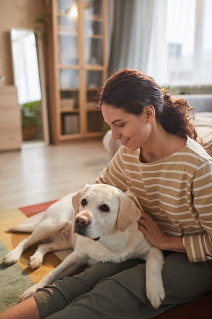 Verticaal warm getinte portret van lachende jonge vrouw die hond knuffelt terwijl ze op de vloer zit in een gezellig interieur