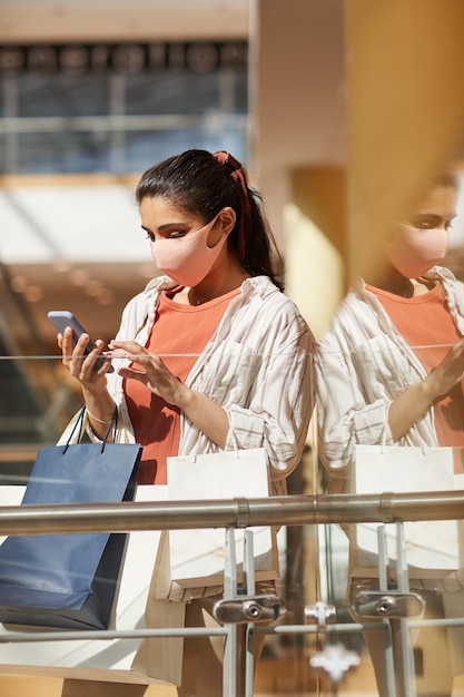 Verticaal zijaanzichtportret van jonge vrouw die masker draagt tijdens het winkelen in wandelgalerij en het gebruik van smartphone, scène verlicht door zonlicht