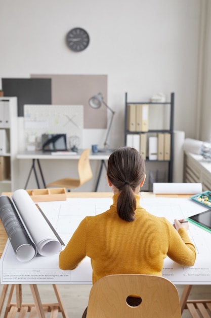 Vertical back view portrait of female architect drawing blueprints while sitting at desk in office