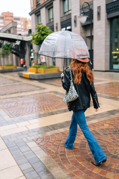 Vertical back view to unrecognizable young woman in fashion hat\
walking on european city street with transparent umbrella enjoying\
rainy weather outdoors