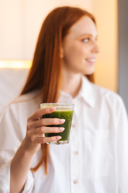Vertical blurred portrait of cheerful attractive young woman holding glass with green vegetable detox smoothie cocktail from blender in kitchen Concept of healthy eating and lifestyle