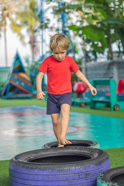 Verticale ragazzo pratica gioco equilibrio coordinazione motoria allenamento salta vai passo su pneumatici per auto concetto divertimento all'aria aperta sport all'aria aperta lezioni di gioco corsa a ostacoli per bambino migliorare l'efficienza alzando il figlio