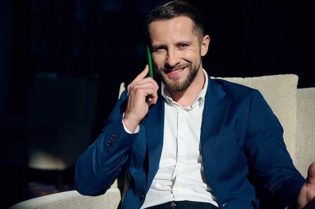 Vertical business portrait of attractive handsome confident Caucasian businessman, entrepreneur talking on mobile phone, smiling confidently, looking at camera, relaxing on an armchair in hotel lobby