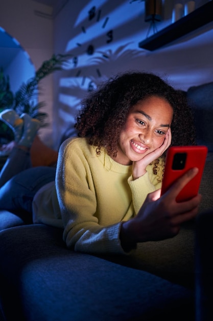 Foto verticale allegra giovane ragazza latina che usa lo smartphone sdraiata sul divano sorridente donna che chatta con il cellulare