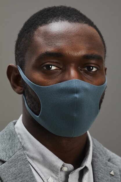 Vertical close up portrait of confident African-American businessman wearing mask and looking at camera while standing against minimal gray background