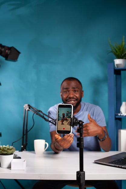 Vertical closeup of smartphone filming influencer sitting down at desk with laptop smiling interacting with fans. Selective focus on live video podcast setup recording content creator talking.