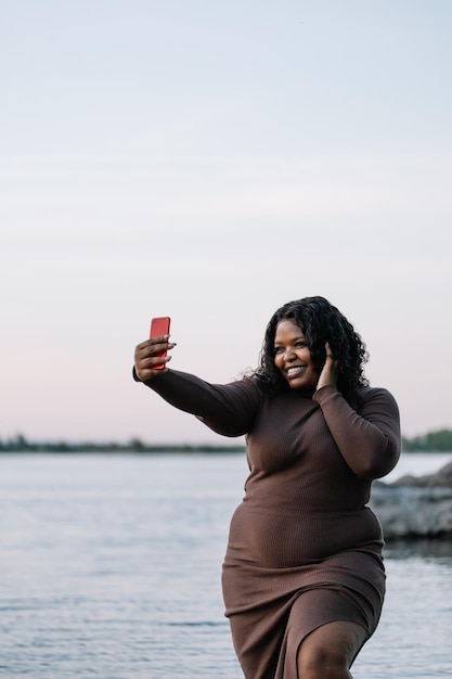 Vertical curly laughing pretty african american woman in elegant dress take selfie by smartphone at sea side Copy space