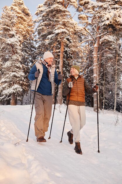 Vertical full length portrait of active senior couple walking\
with poles in beautiful winter forest