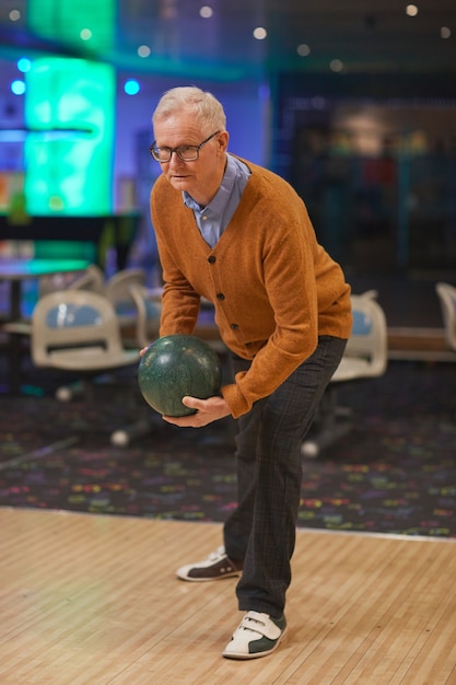 Foto ritratto verticale a figura intera di un uomo anziano attivo che gioca a bowling, in piedi vicino alla corsia pronto a lanciare mentre si gode l'intrattenimento