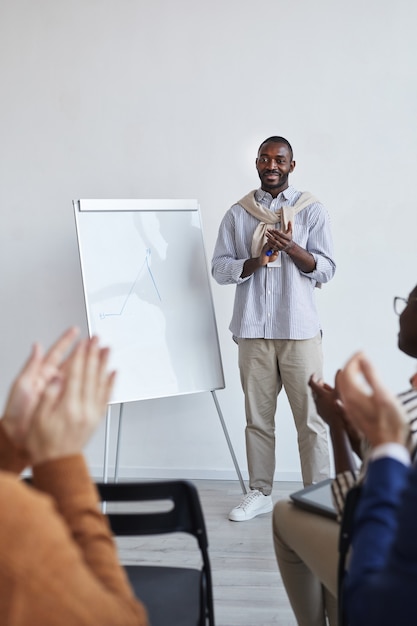 Ritratto verticale a figura intera di business coach afro-americano che parla con il pubblico a una conferenza o a un seminario educativo mentre è in piedi accanto alla lavagna e applaude