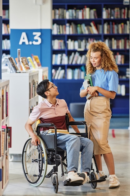 Вертикальный портрет азиатского студента с инвалидностью в полный рост, разговаривающего с другом в библиотеке колледжа