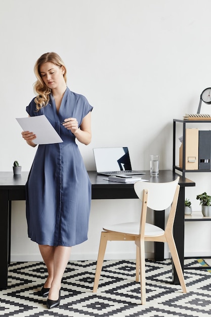 Vertical full length portrait of confident blonde businesswoman wearing dress while standing by desk...