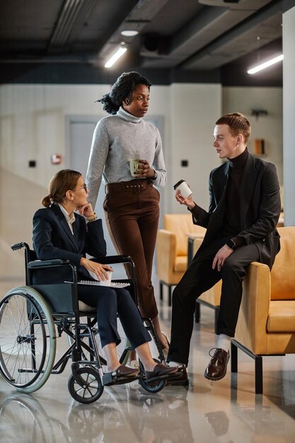 Vertical full length portrait of diverse business team with young woman in wheelchair chatting to colleagues in office